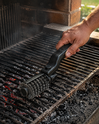 Mano usando el cepillo eléctrico sobre la parrilla de un quincho con brasas encendidas